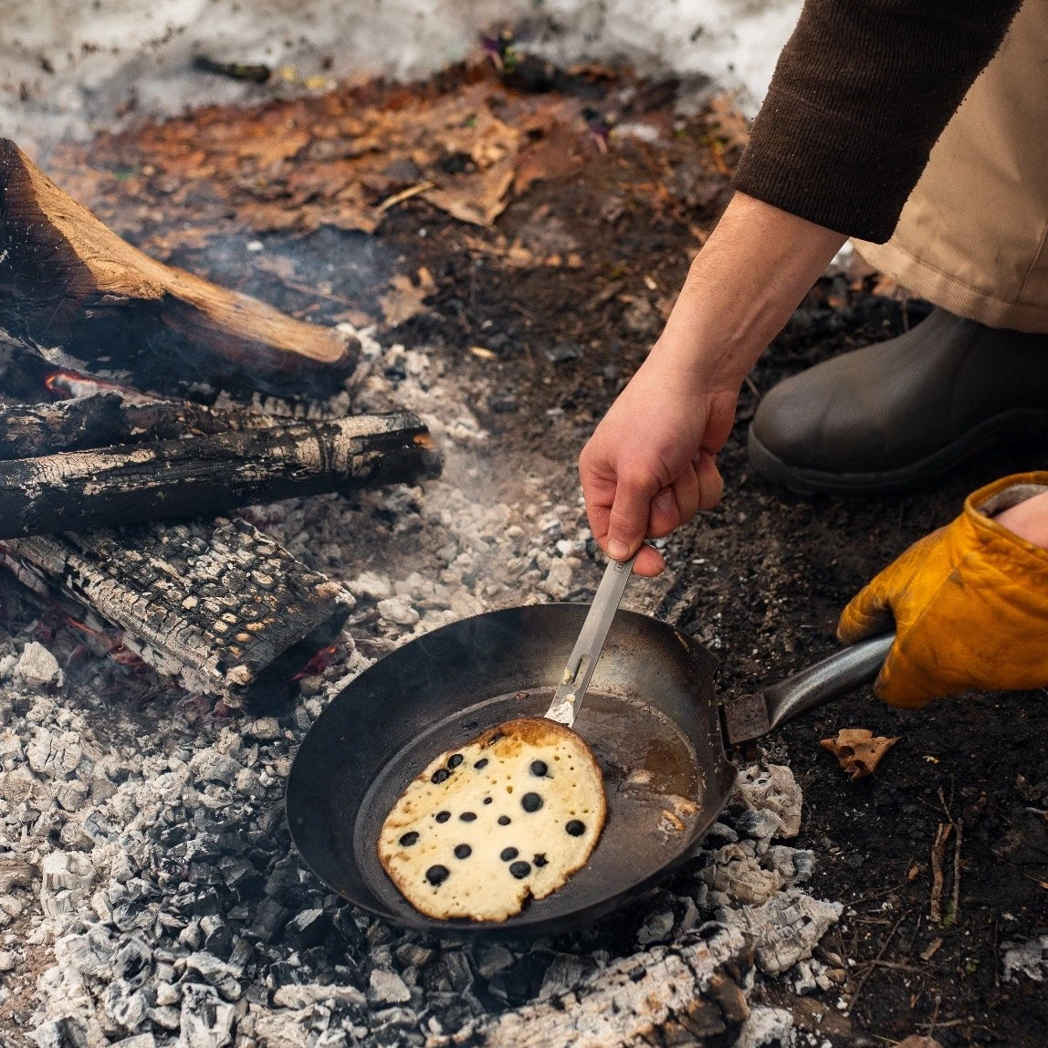 A blueberry pancake cooking in a skillet over campfire coals