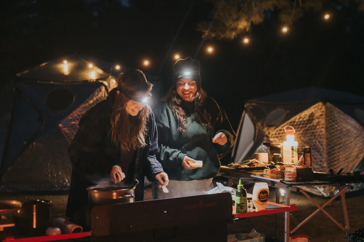 Friends cooking together at a campsite under evening string lights