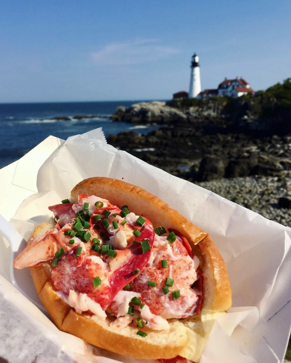 Bite Into Maine lobster roll with Portland Head Light behind it