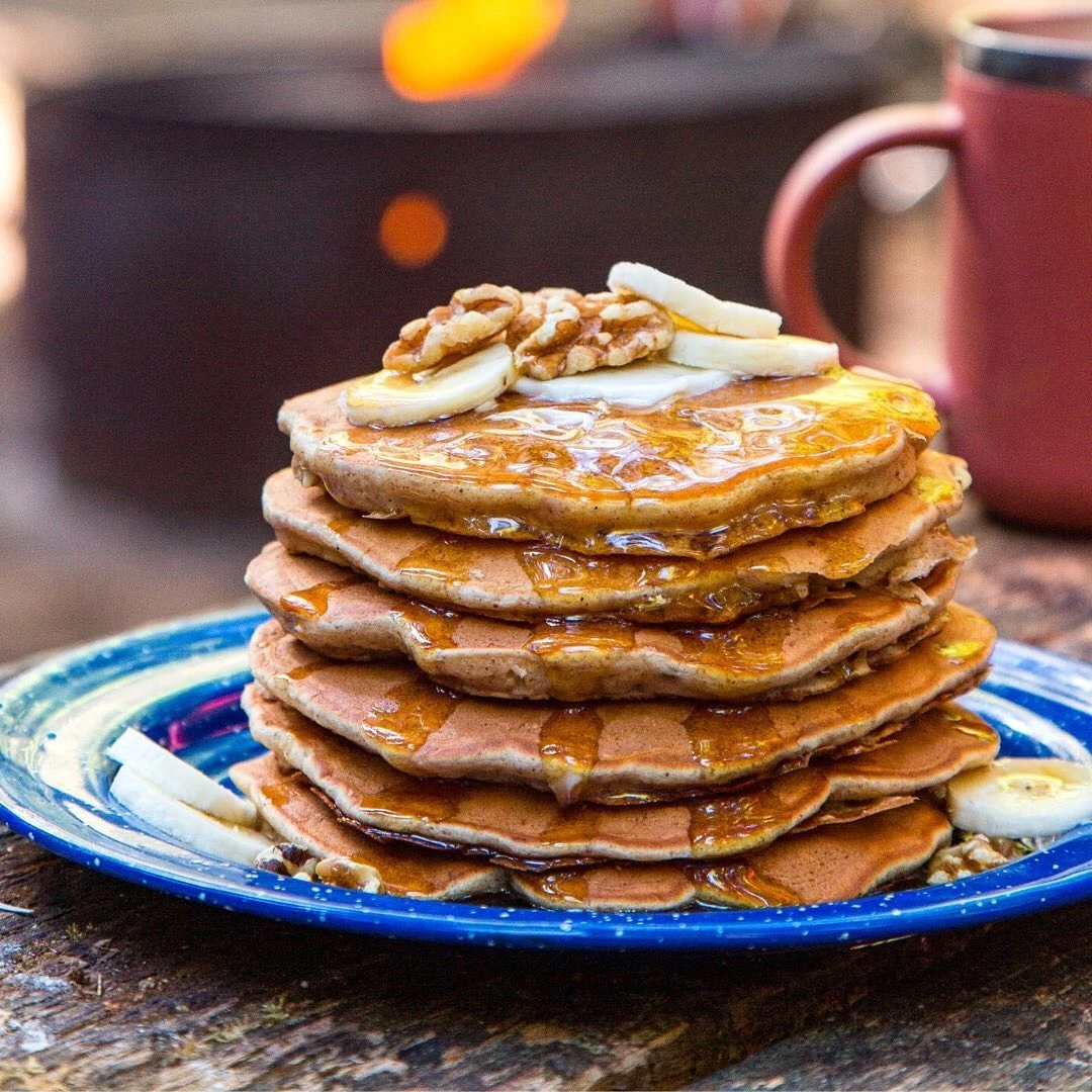 Banana pancakes topped with banana slices, walnuts, and syrup beside a campfire